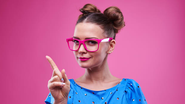 woman in a blue dress and pink glasses holds up her index finger. .